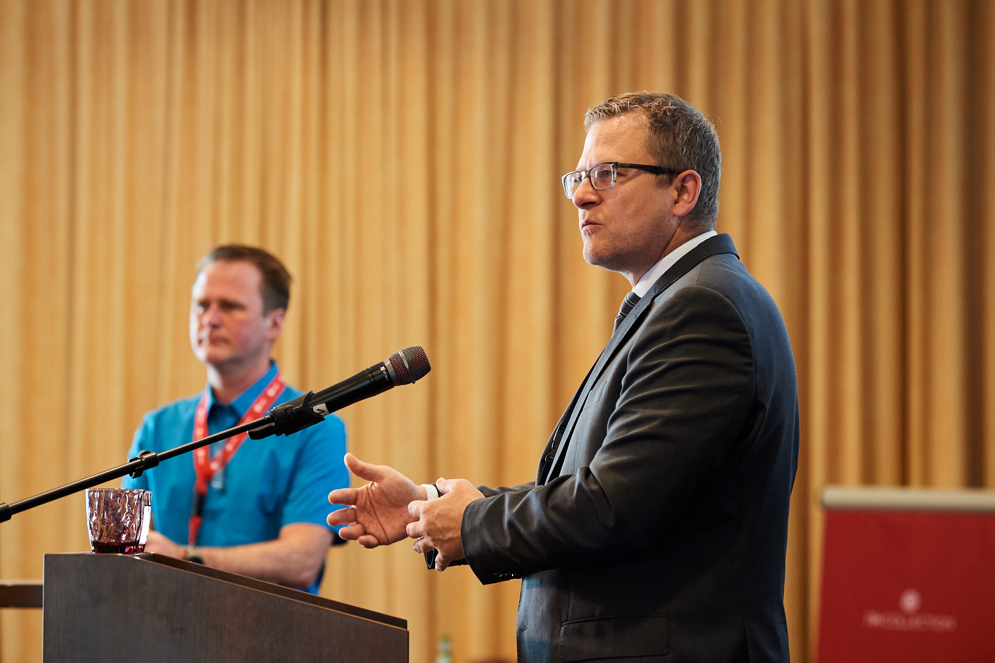 Zwei Männer befinden sich auf der Bühne der Konferenz, einer spricht ins Mikrofon.