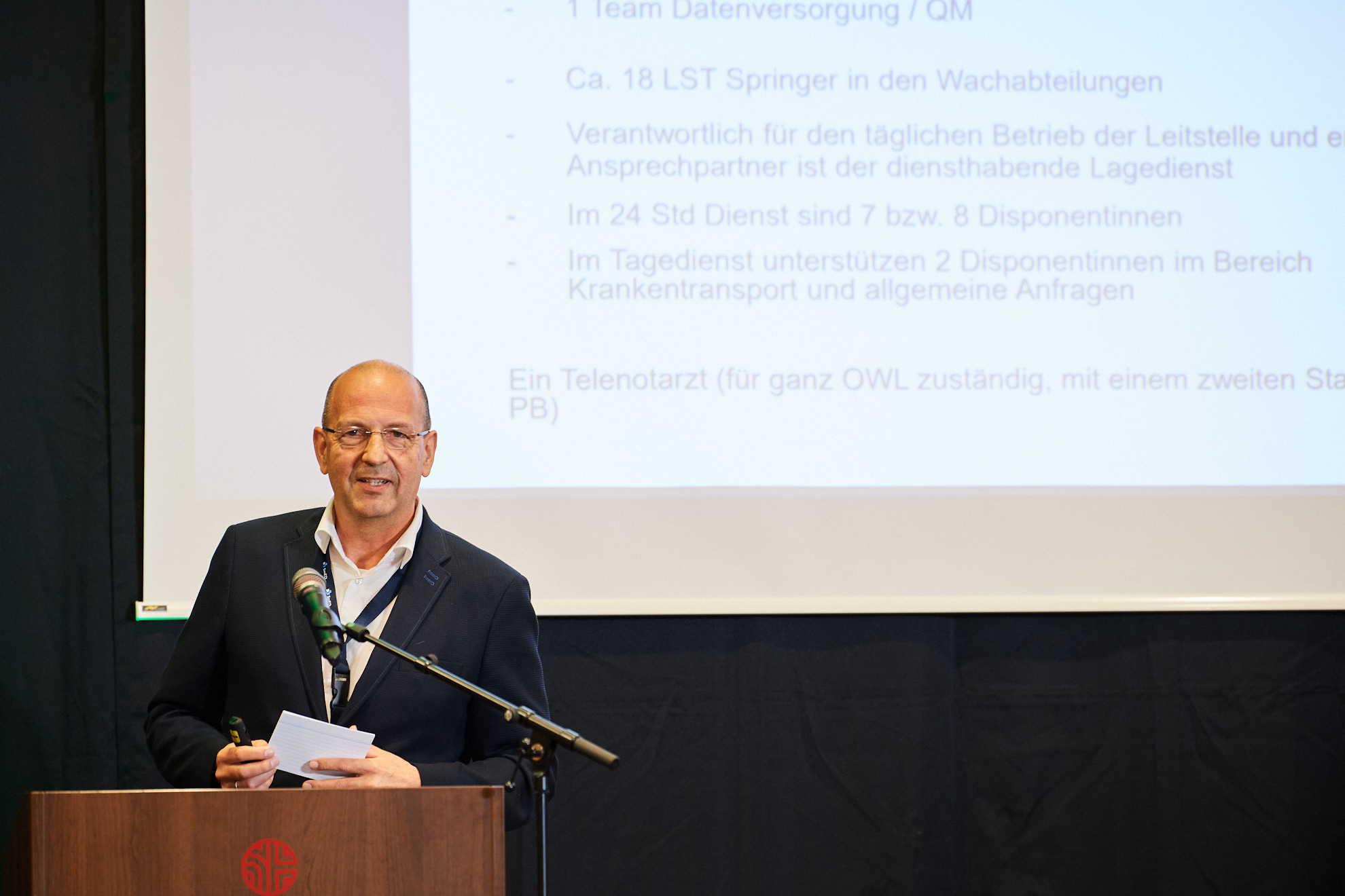 Ein Mann hält einen Vortrag, im Hintergrund ist die Präsentation auf die Wand projiziert.