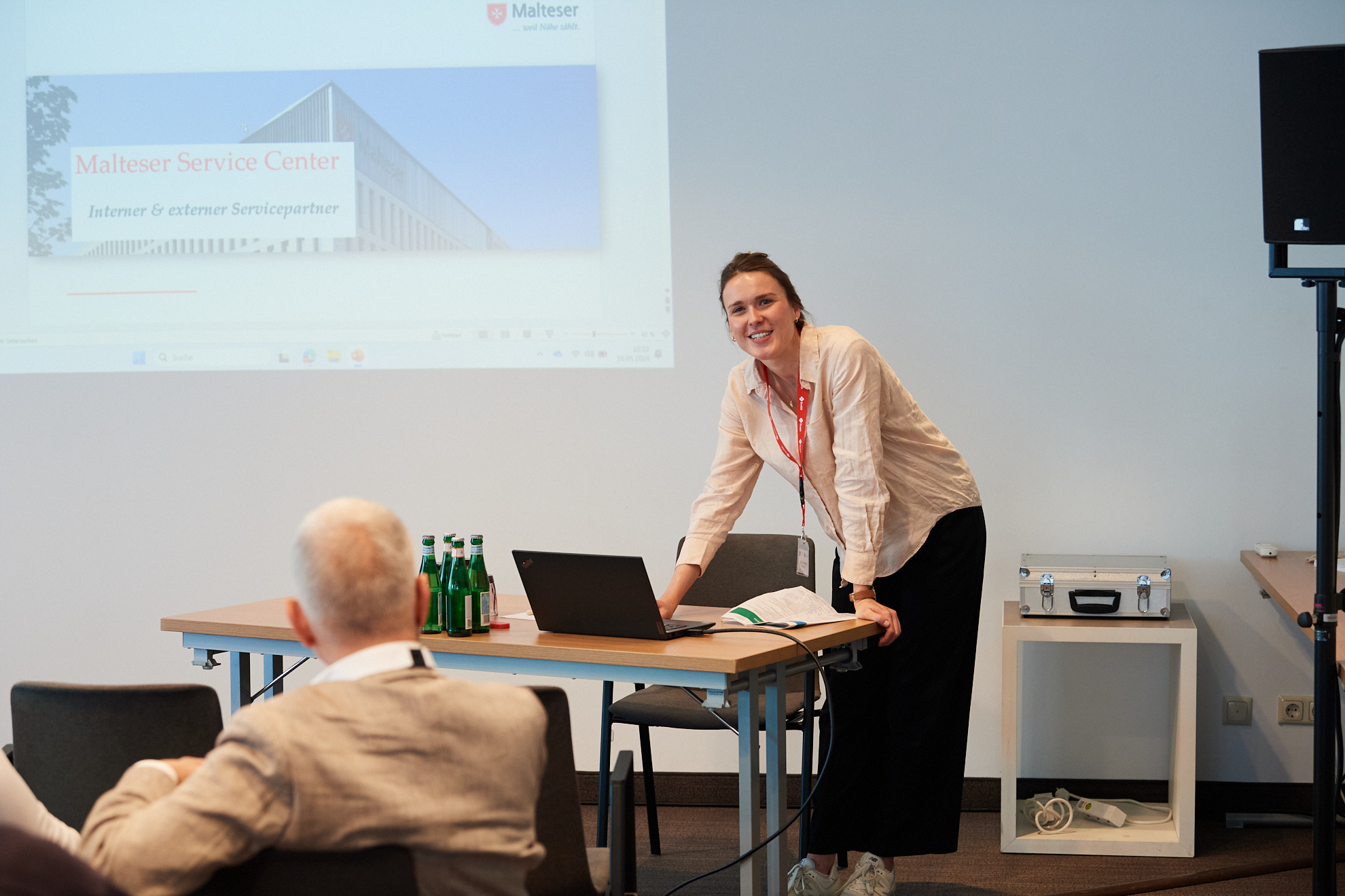 Eine Frau steht lächelnd neben einem Tisch mit einer Hand auf einem Laptop und hält eine Präsentation.