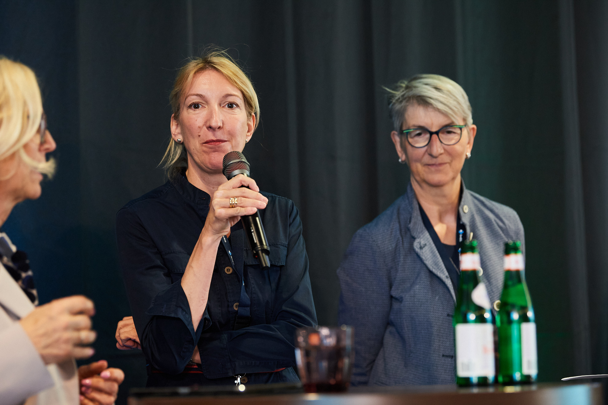 Drei Frauen sind auf der Bühne, eine von ihnen spricht in ein Mikrofon.
