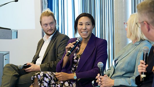 Podiumsteilnehmende während ihres Wortbeitrags neben weiteren Podiumsgästen