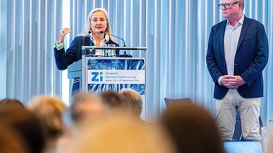 Rednerin am Podium während eines Vortrags. Zweiter Redner steht ihr zur Seite.